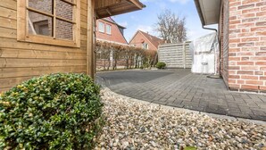 Een houten huis met een verharde oprit, een bakstenen muur en een kleine tuin met grind en struiken.