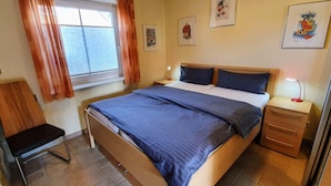 Een slaapkamer met een bed, nachtkastjes, een stoel, een raam met gordijnen en kunst aan de muren.