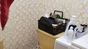 Une salle de bain avec des toilettes, un lavabo et un meuble. On trouve un panier sur le couvercle des toilettes et divers articles de toilette sur le comptoir.