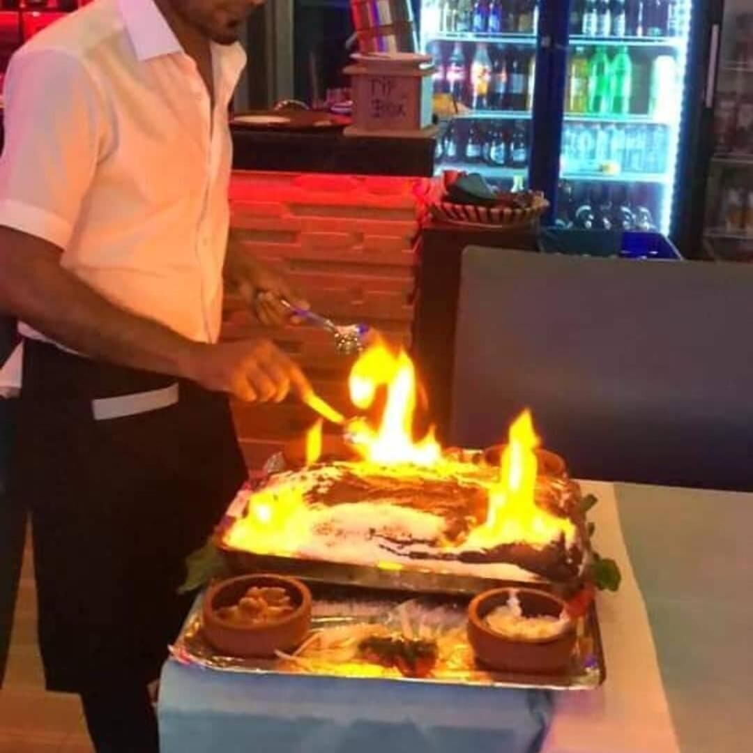 A person lighting a large, rectangular cake with flames on a table.