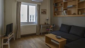 Uma sala de estar com sofá, mesa de centro, televisão sobre um suporte, estante de livros e janela com cortinas.