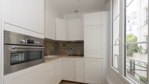 Cocina compacta con gabinetes blancos, horno de acero inoxidable, salpicadero decorativo y ventana con vistas al exterior.
