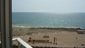 A beach view from a balcony with a clear sky, a sandy beach, and a parking area with a few cars and benches.