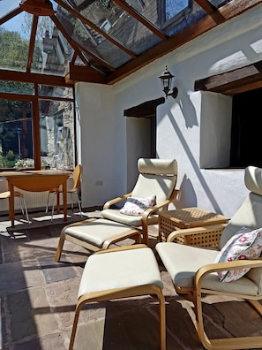 A sunlit patio with wicker lounge chairs and a table, a stone floor, and a view of a stone building through large windows. - Perfect Rural Retreat for two near Hay on Wye (Hereford)