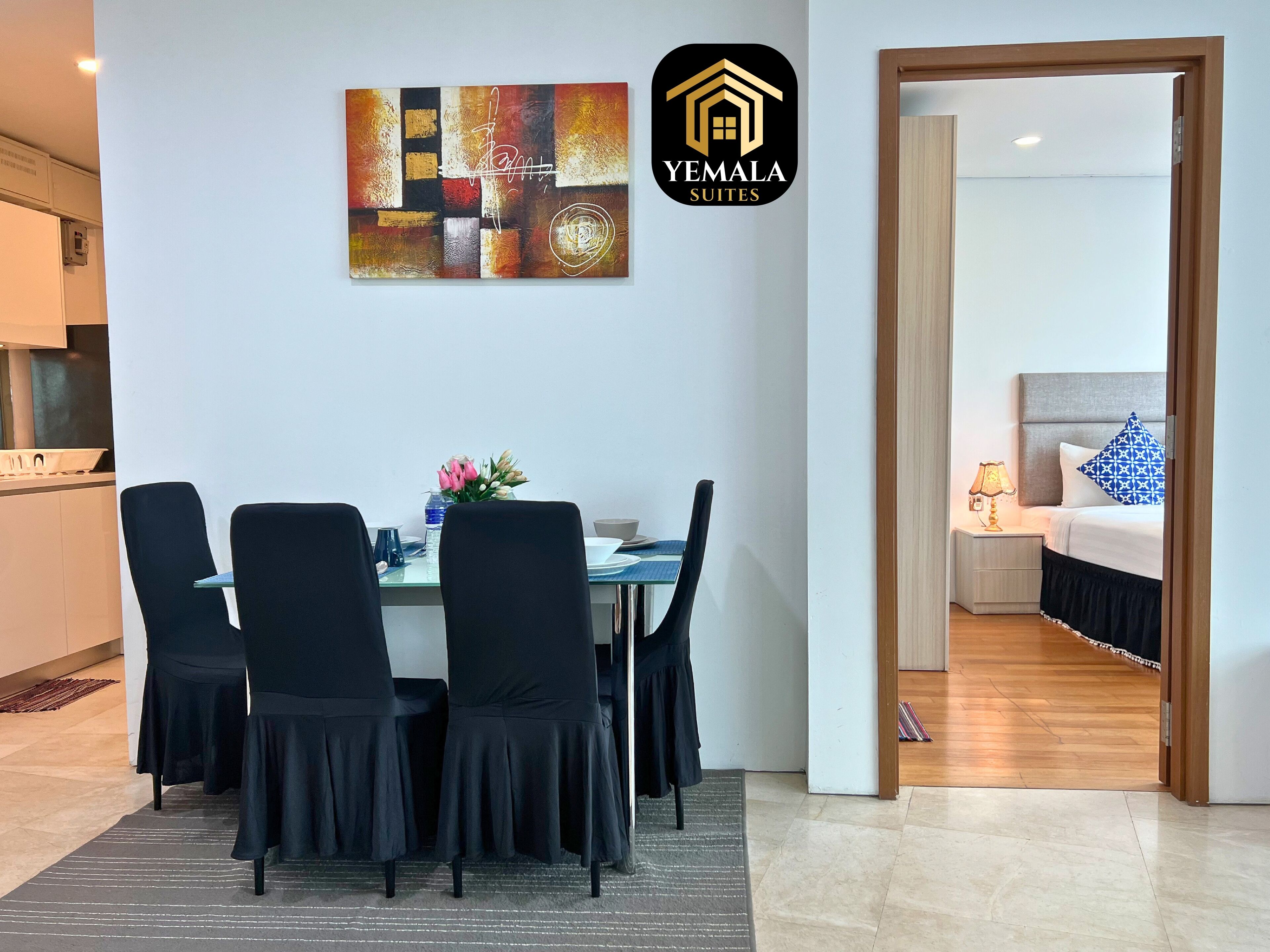 A modern dining area with a table set for four, black chairs, and a vase of flowers. A logo for 'Yemala Suites' is visible on the wall.