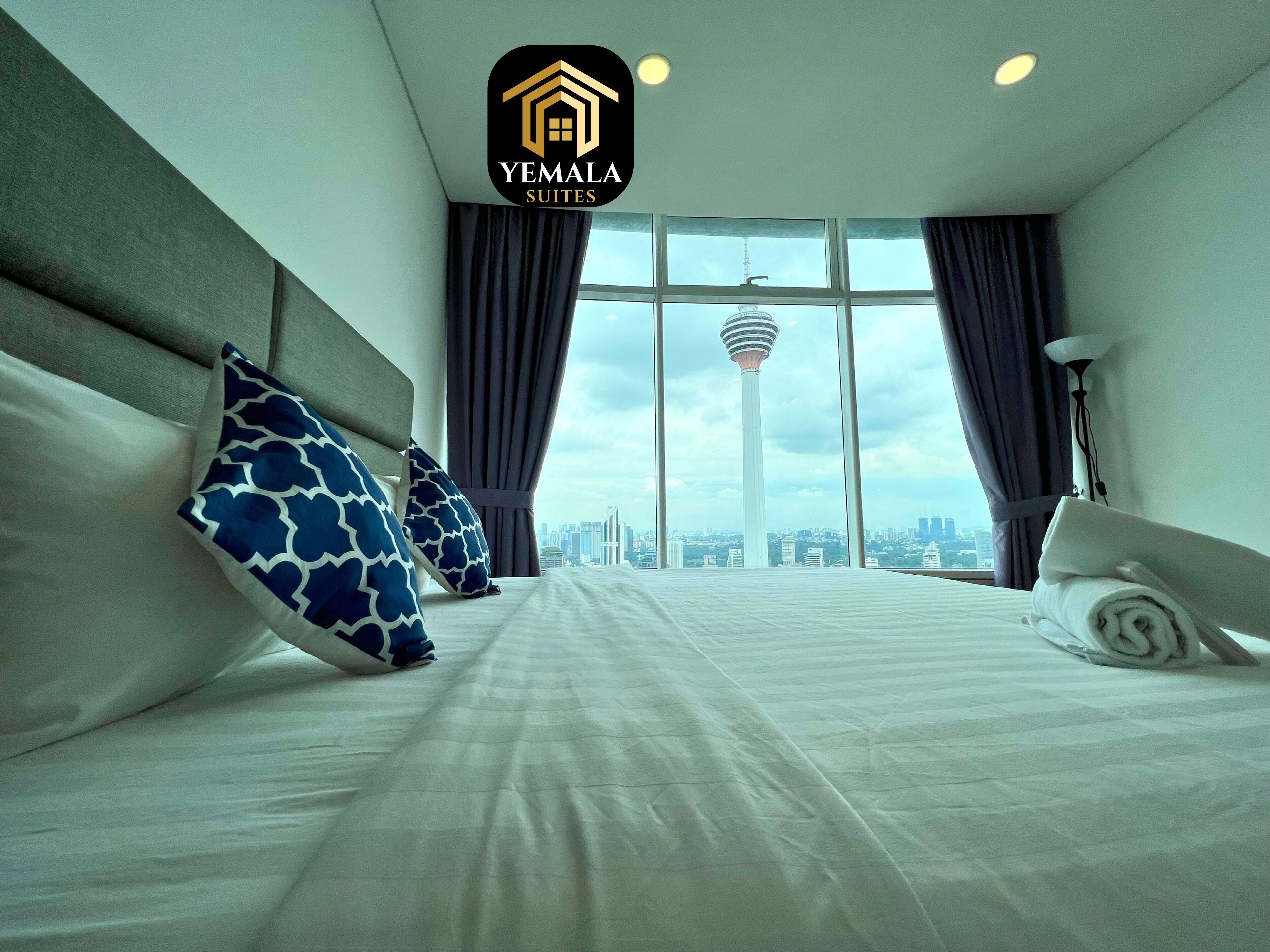 A hotel room with a large bed, two pillows, a nightstand, a lamp, and a view of a cityscape and tower.