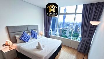 Habitación de hotel con una cama grande, una mesita de noche con lámpara, vistas de la ciudad a través de amplios ventanales y el logotipo de Yemala Suites.