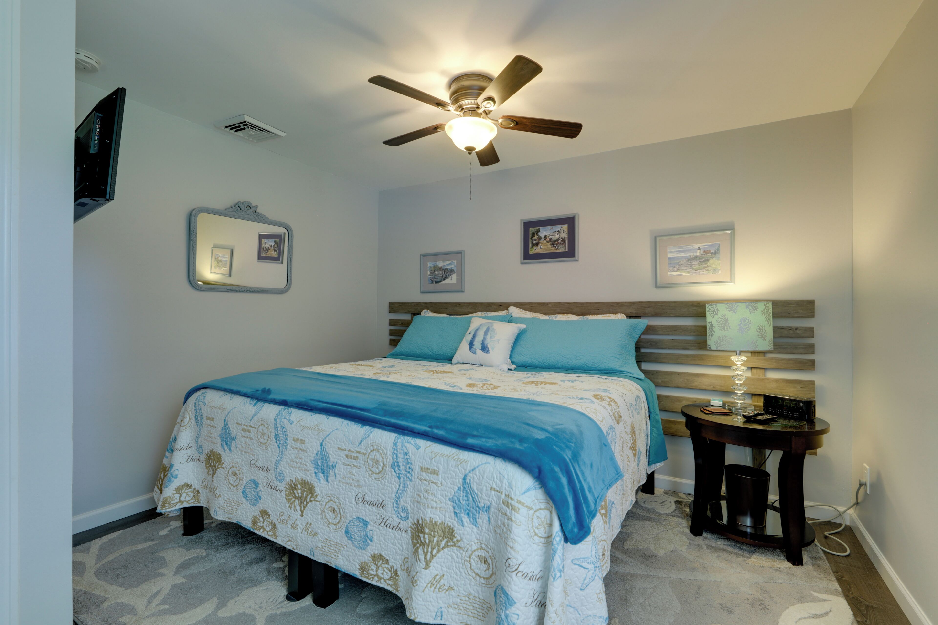 A bedroom with a bed, a ceiling fan, a bedside table with a lamp, and framed pictures on the wall.