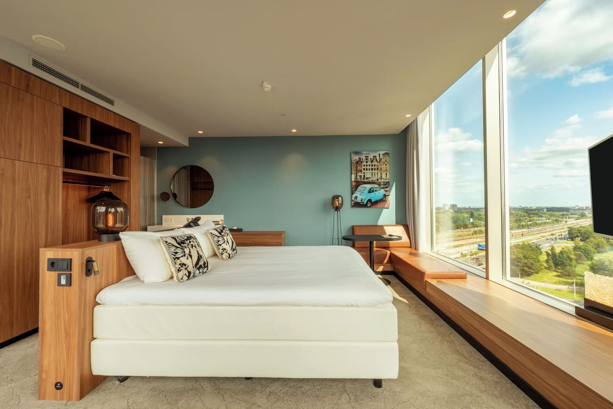 Een moderne slaapkamer met een groot bed, een inbouwkast van hout, een ronde spiegel en uitzicht op een stadsbeeld door grote ramen.