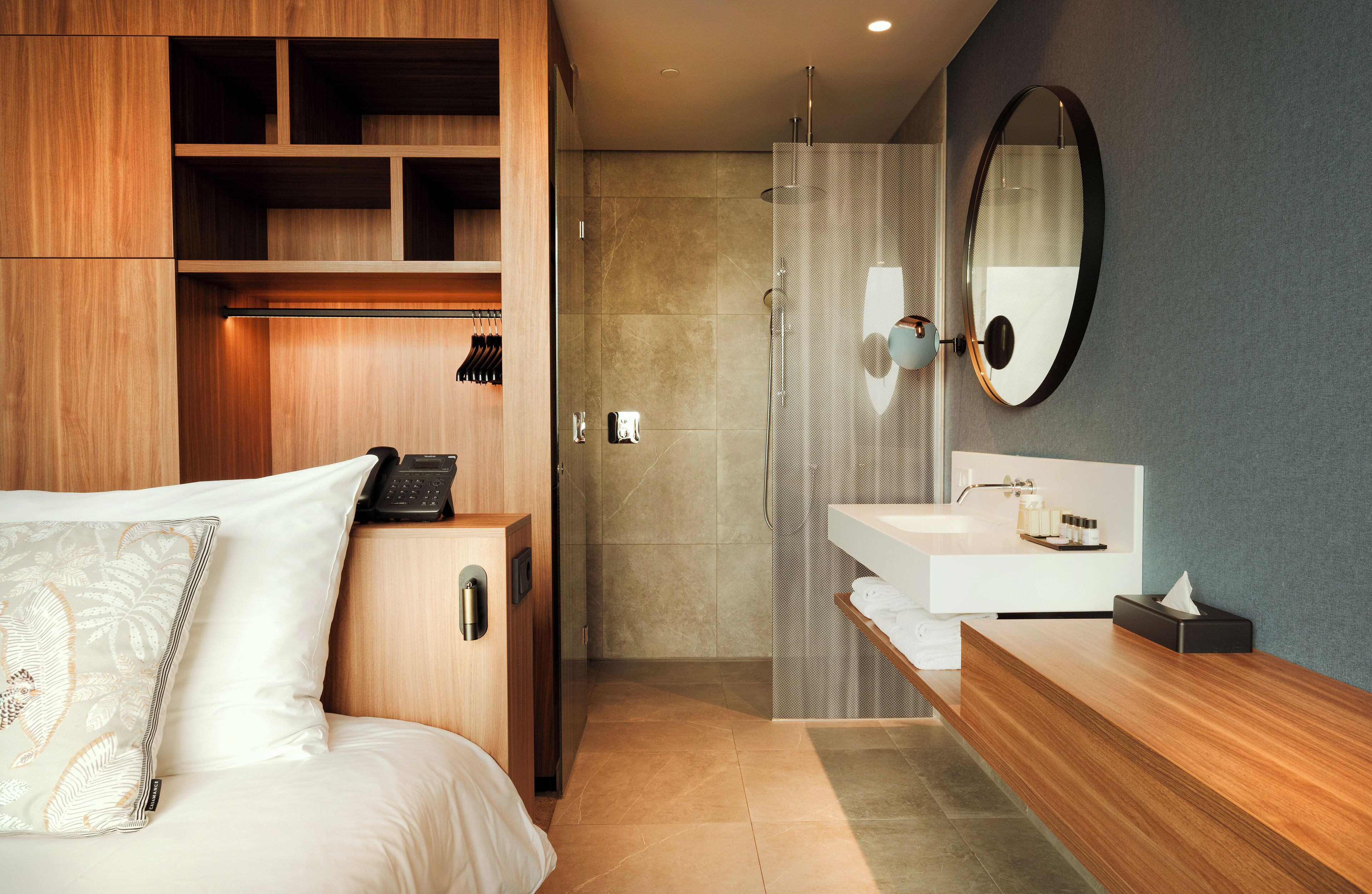 Quarto de hotel moderno com guarda-roupa de madeira, cama com almofada decorativa, banheiro com espelho grande, chuveiro e toucador branco com artigos de higiene pessoal.