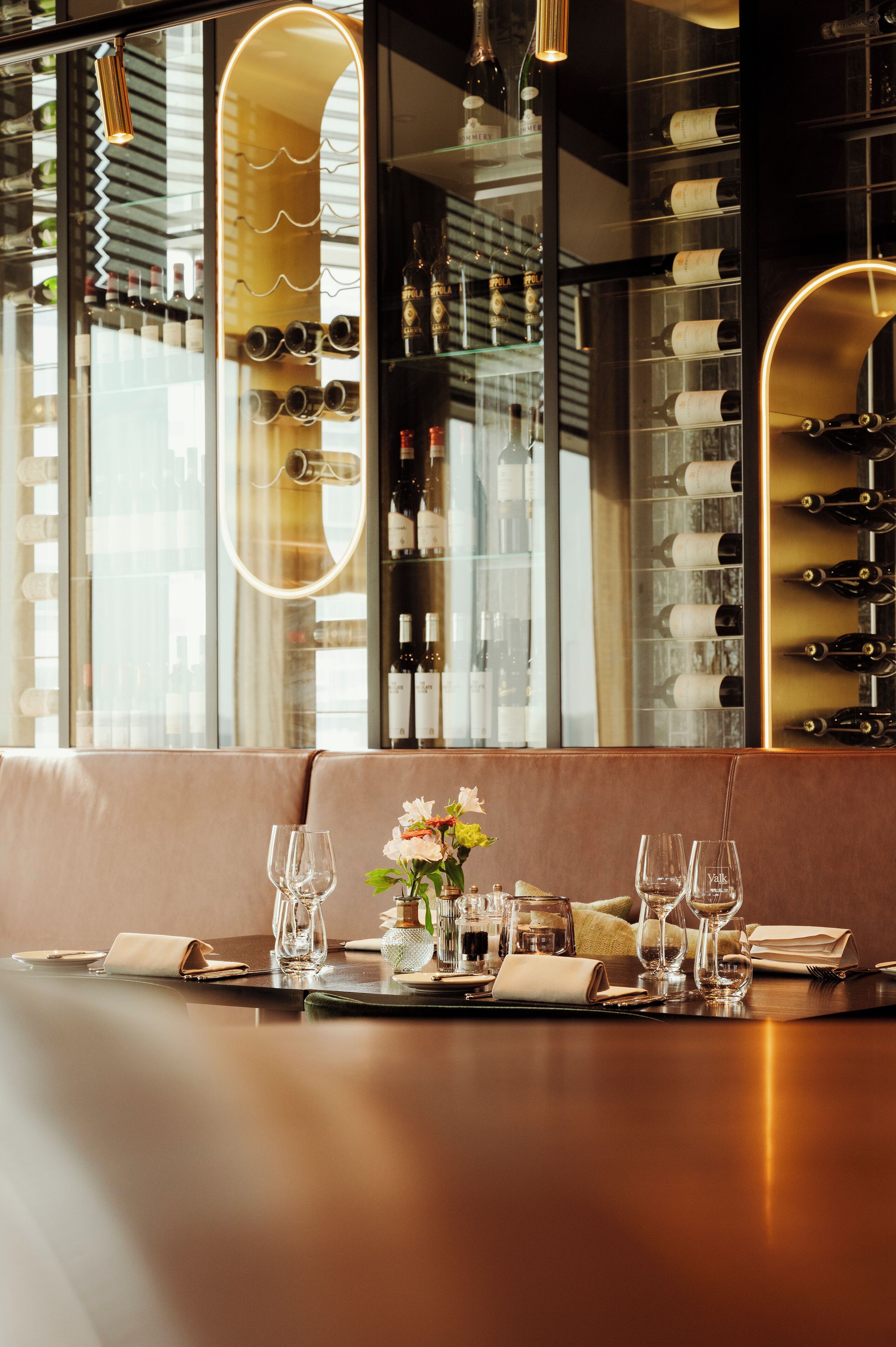 A restaurant interior with a long table set for dining, a wine rack in the background, and a large window offering a view.