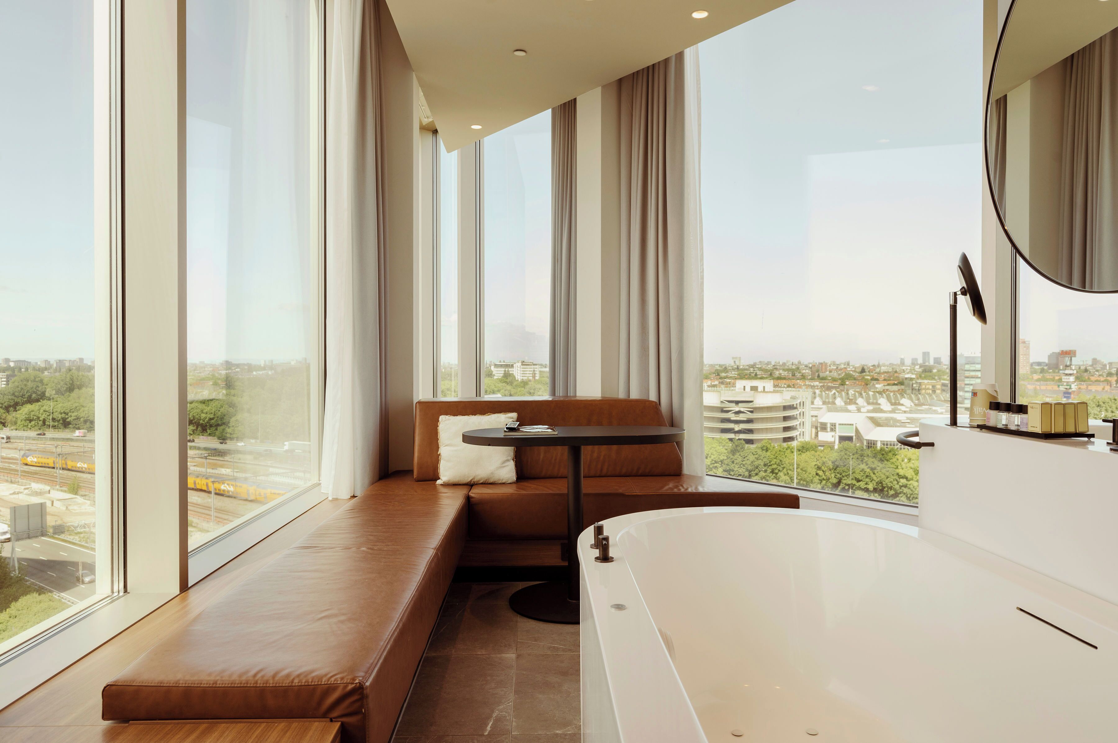 A modern bathroom with a large bathtub, a seating area with a table, and a view of the city through large windows.