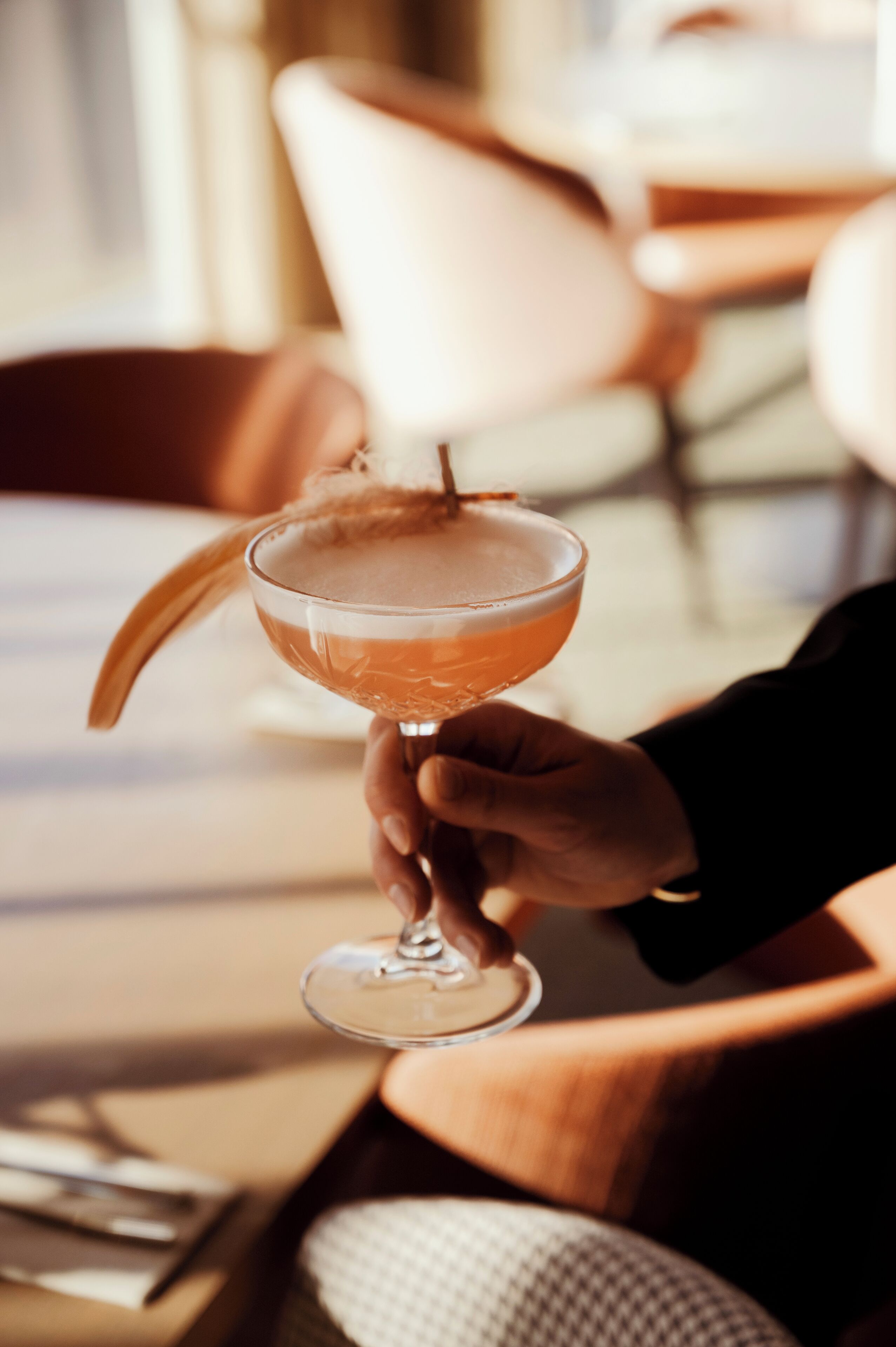 Une personne tenant un verre à cocktail orné d’une garniture incurvée, assise à une table, avec une chaise visible à l’arrière-plan.