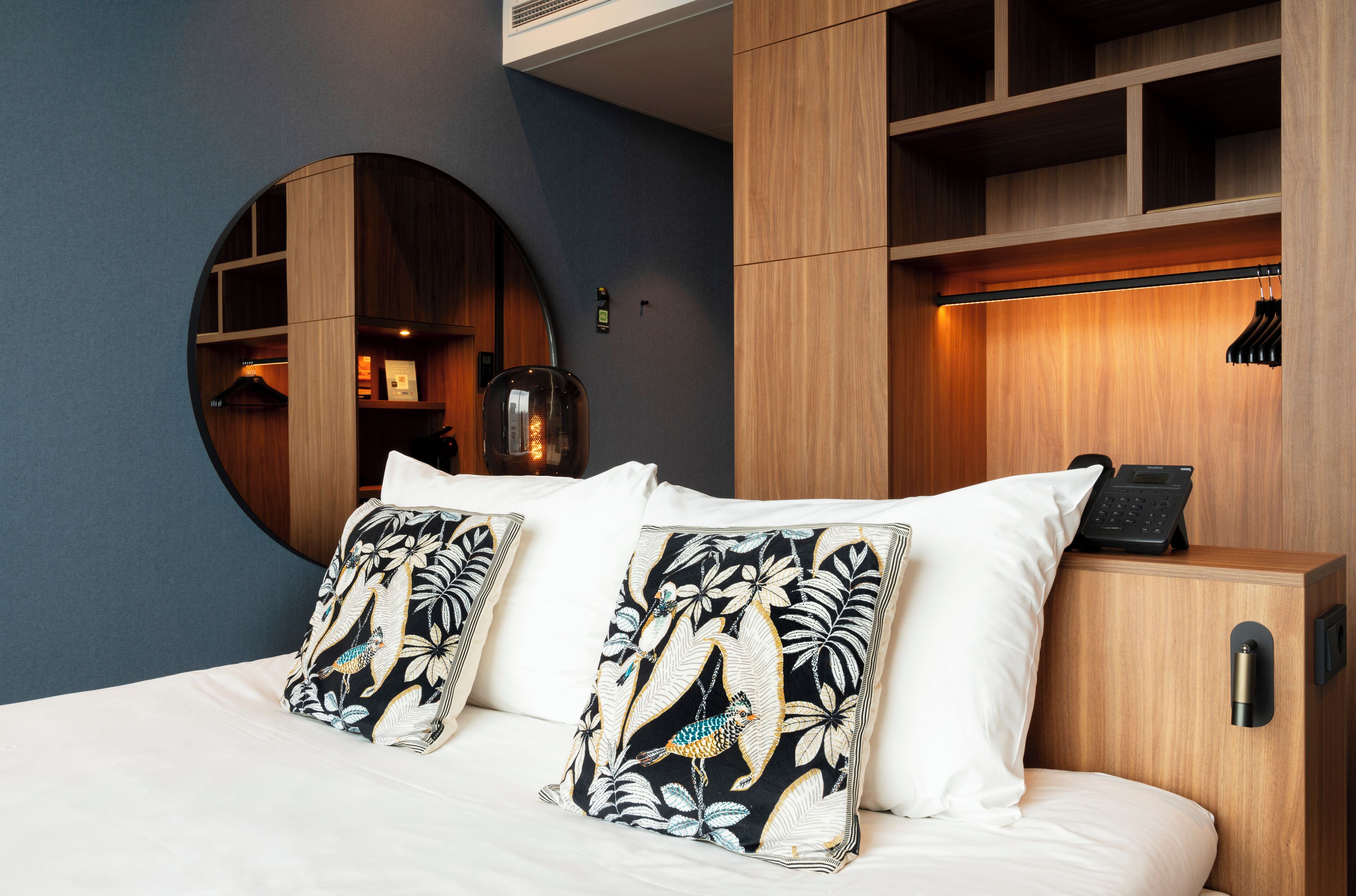 A hotel room with a bed, two pillows with a bird and leaf pattern, a round mirror, and a wooden wardrobe with a hanging rack.