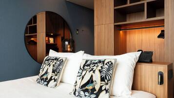A hotel room with a bed, two pillows with a bird and leaf pattern, a round mirror, and a wooden wardrobe with a hanging rack.