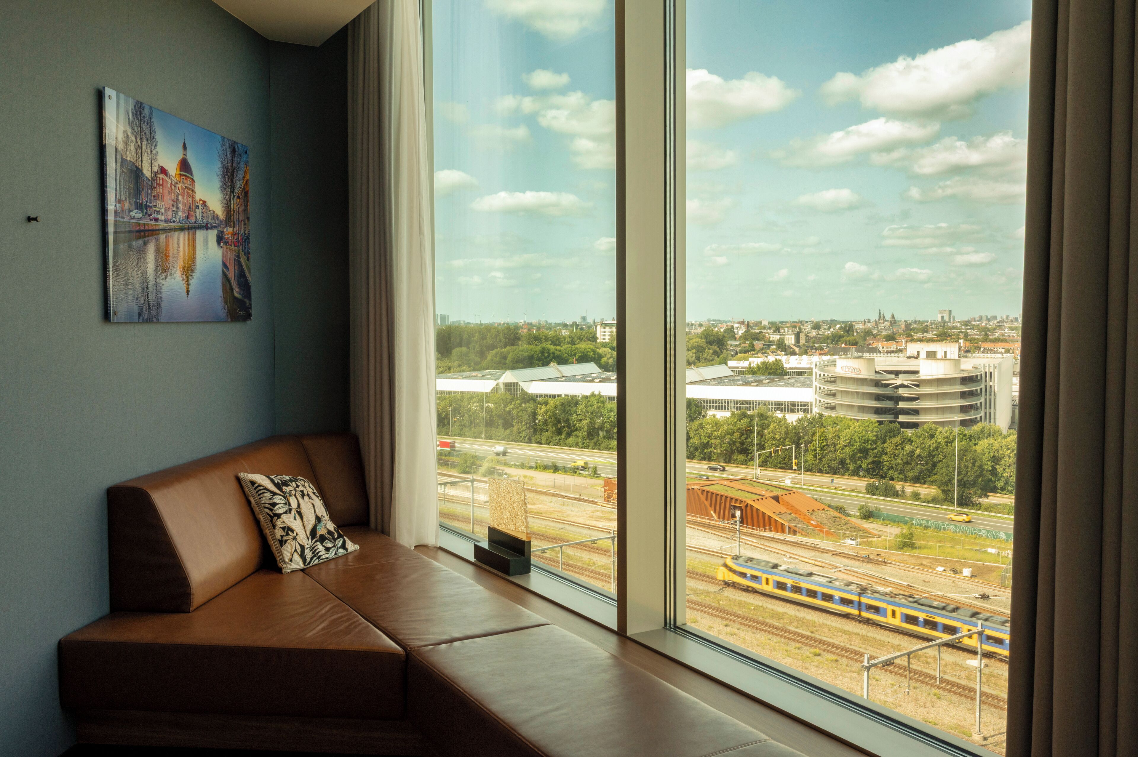 Una sala de estar moderna con un amplio ventanal que ofrece vistas de la ciudad, un sofá de cuero marrón y un cuadro en la pared.