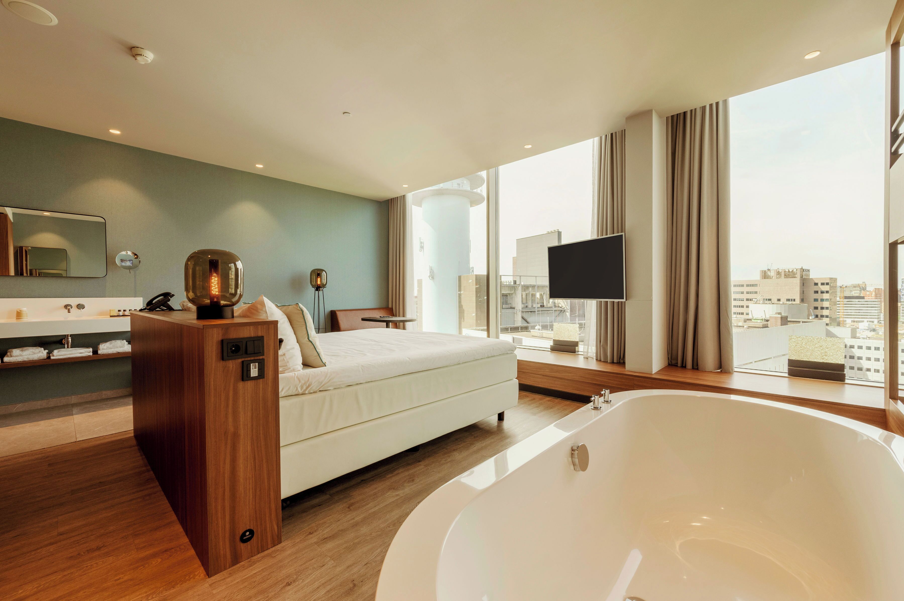 Une chambre d’hôtel moderne dotée d’une grande baignoire, d’un lit, d’une coiffeuse avec un miroir et offrant une vue sur la ville grâce à de grandes fenêtres.