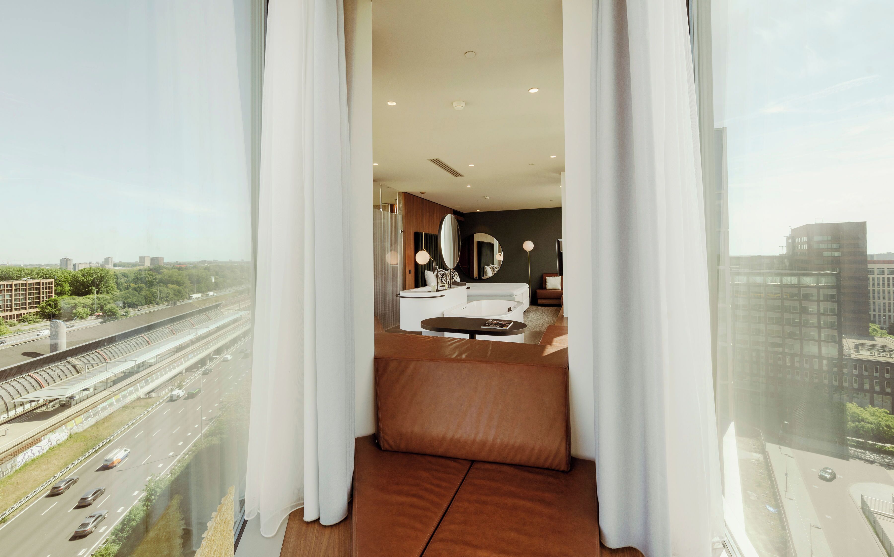 Un dormitorio moderno con una ventana grande, una cama, un tocador y vistas a una autopista y edificios.