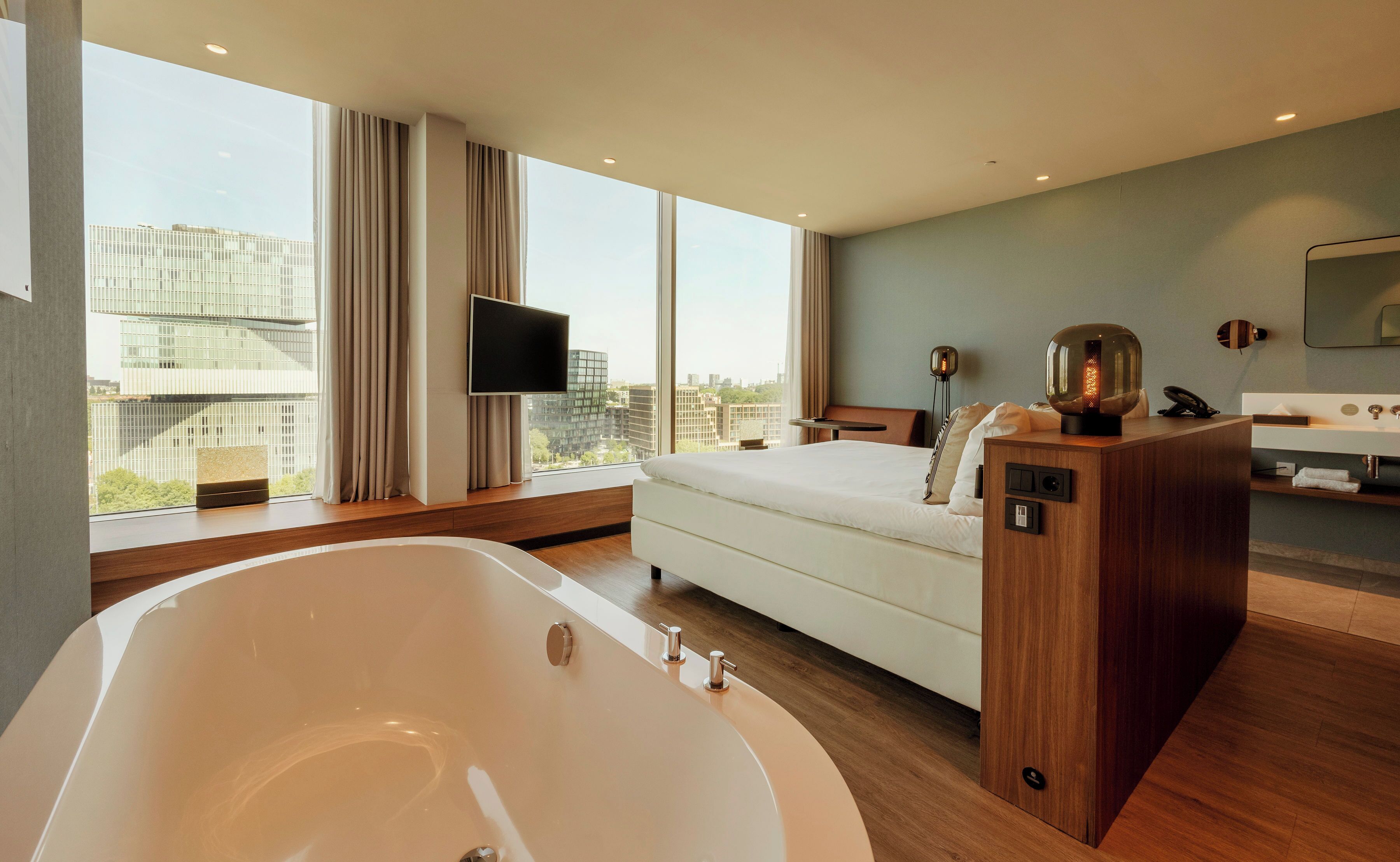 A modern hotel room with a large bathtub, a bed, a TV, and a view of the city.