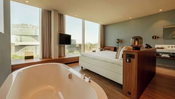 A modern hotel room with a large bathtub, a bed, a TV, and a view of the city.