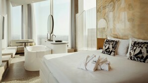 Une chambre d’hôtel moderne dotée d’un grand lit, d’un coin salon avec un canapé et une table ronde, d’une baignoire et d’une grande fenêtre offrant une vue panoramique.