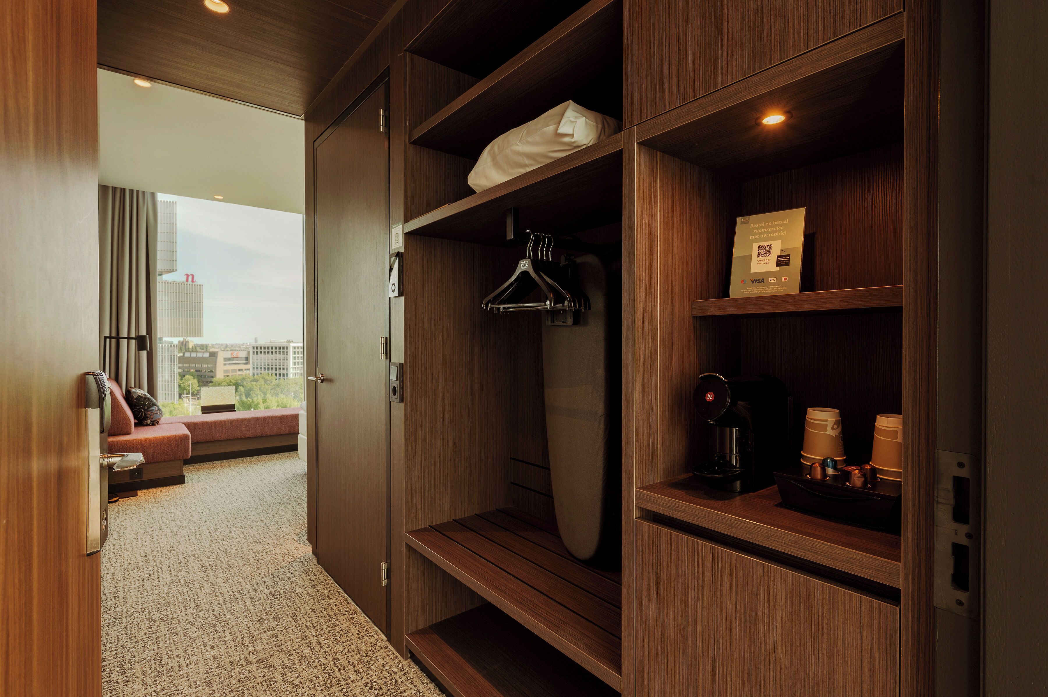 A hotel room with a wooden wardrobe, a hanging garment, a mirror, a coffee maker, and a view of the city.