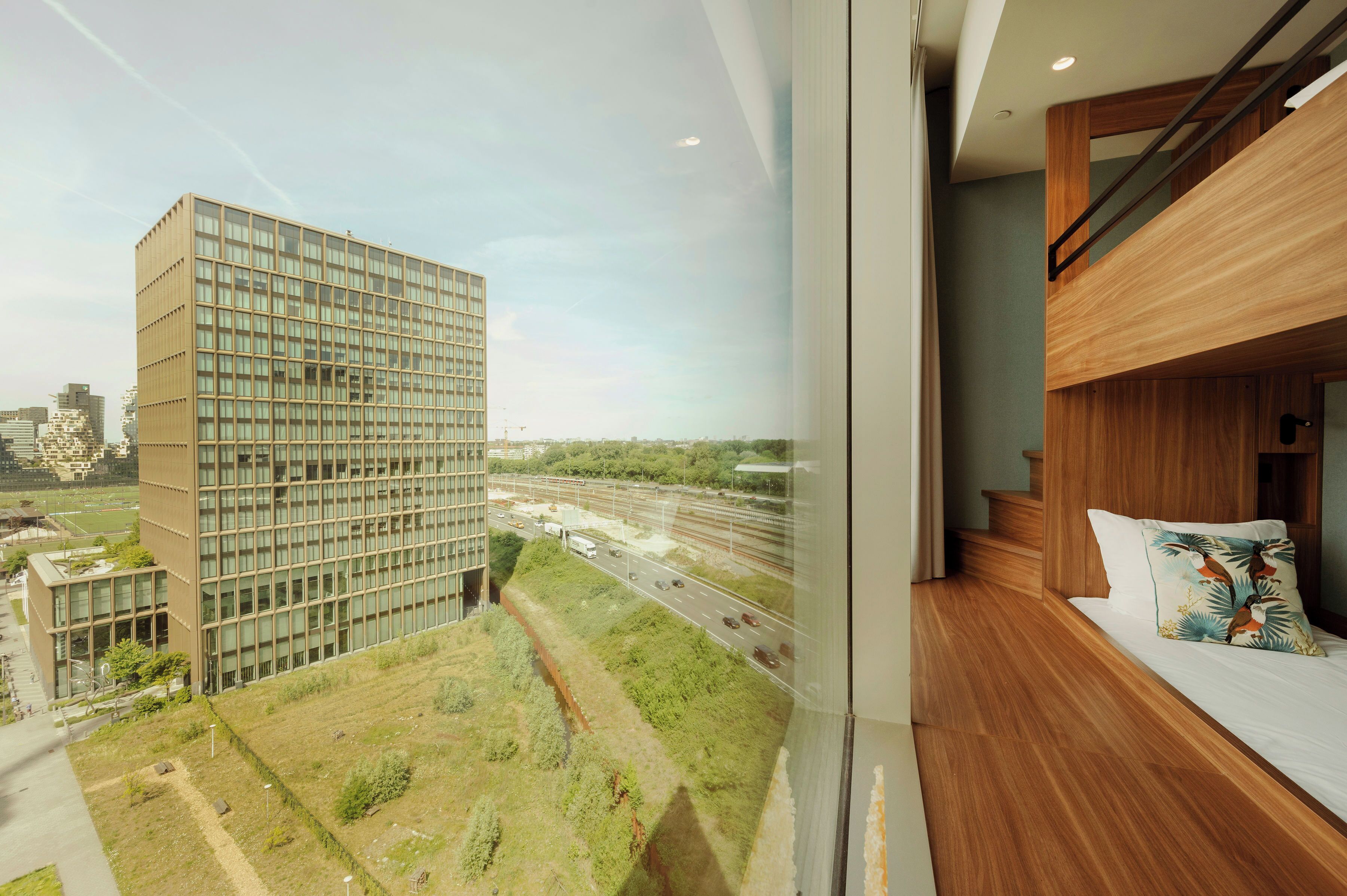 A high-rise building with a modern glass facade, a view of a highway, and a bedroom with a wooden headboard and a patterned pillow.