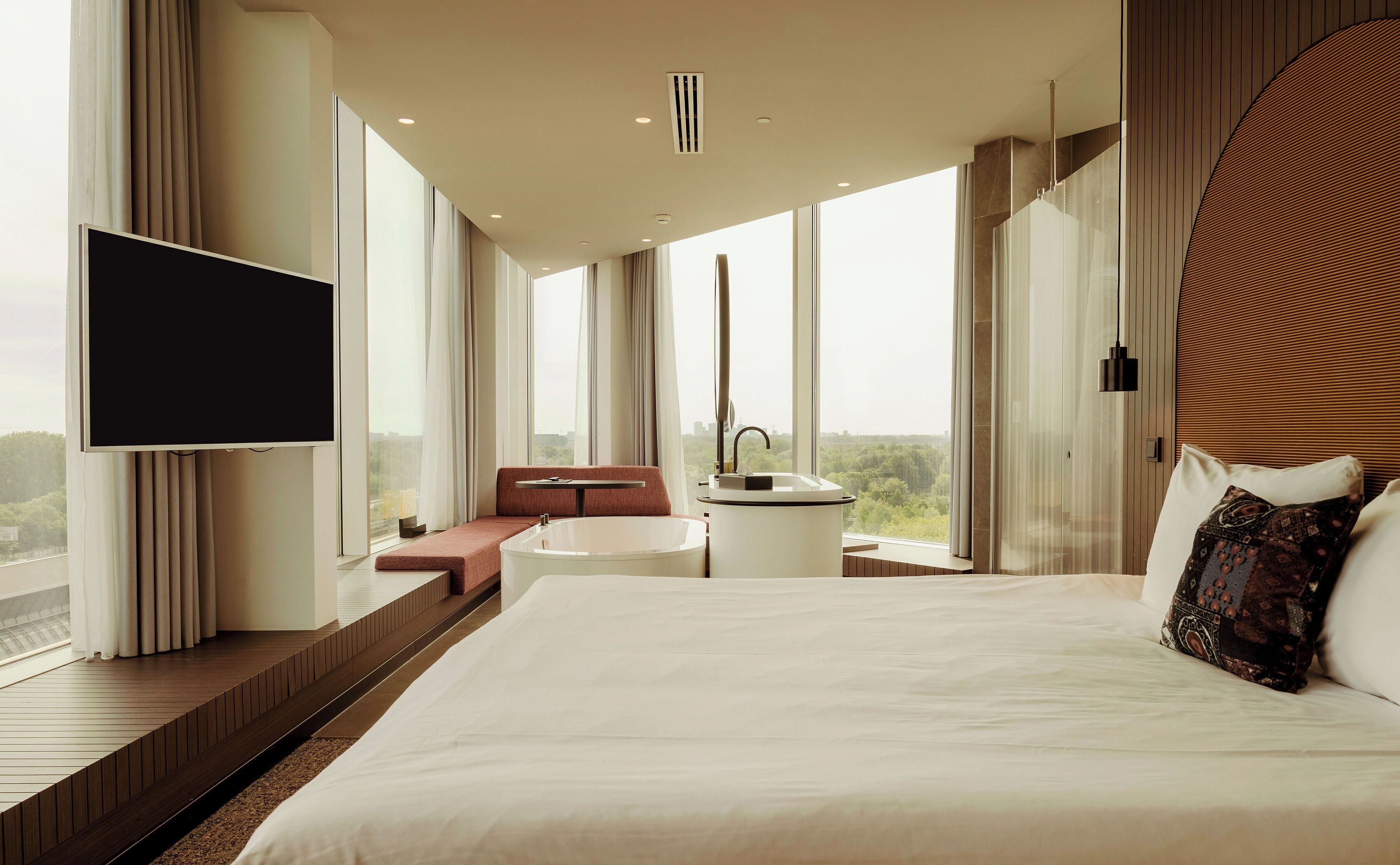 Een moderne hotelkamer met een groot bed, een aan de muur bevestigde televisie, een badkamer met wastafel en spiegel, en uitzicht op de stad door grote ramen.