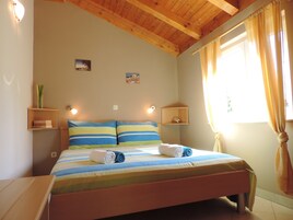 Quarto com cama, cabeceira de madeira, duas mesas de cabeceira, luminárias na parede, uma janela com cortinas e um pequeno prateleira com uma planta.