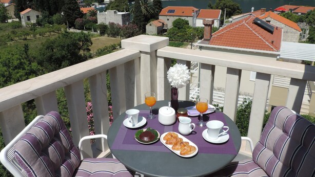 A balcony with a table set for breakfast, including croissants and orange juice, overlooking a scenic village with houses and a river. - Apartments Vodnica (Dubrovnik)