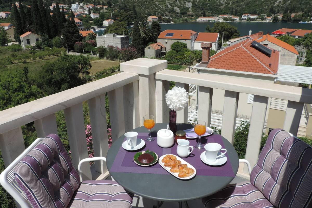Un balcon avec une table dressée pour le petit-déjeuner, comprenant des croissants et du jus d’orange, donnant sur un village pittoresque avec ses maisons et sa rivière.