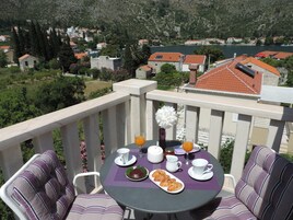 Varanda com mesa posta para o café da manhã, incluindo croissants e suco de laranja, com vista para uma vila pitoresca com casas e um rio.