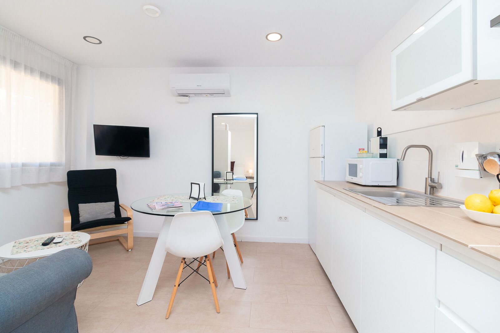 Una cucina moderna con mobili bianchi, un forno a microonde e un lavandino. Si nota un tavolo da pranzo rotondo con sedie e un televisore a schermo piatto montato a parete.
