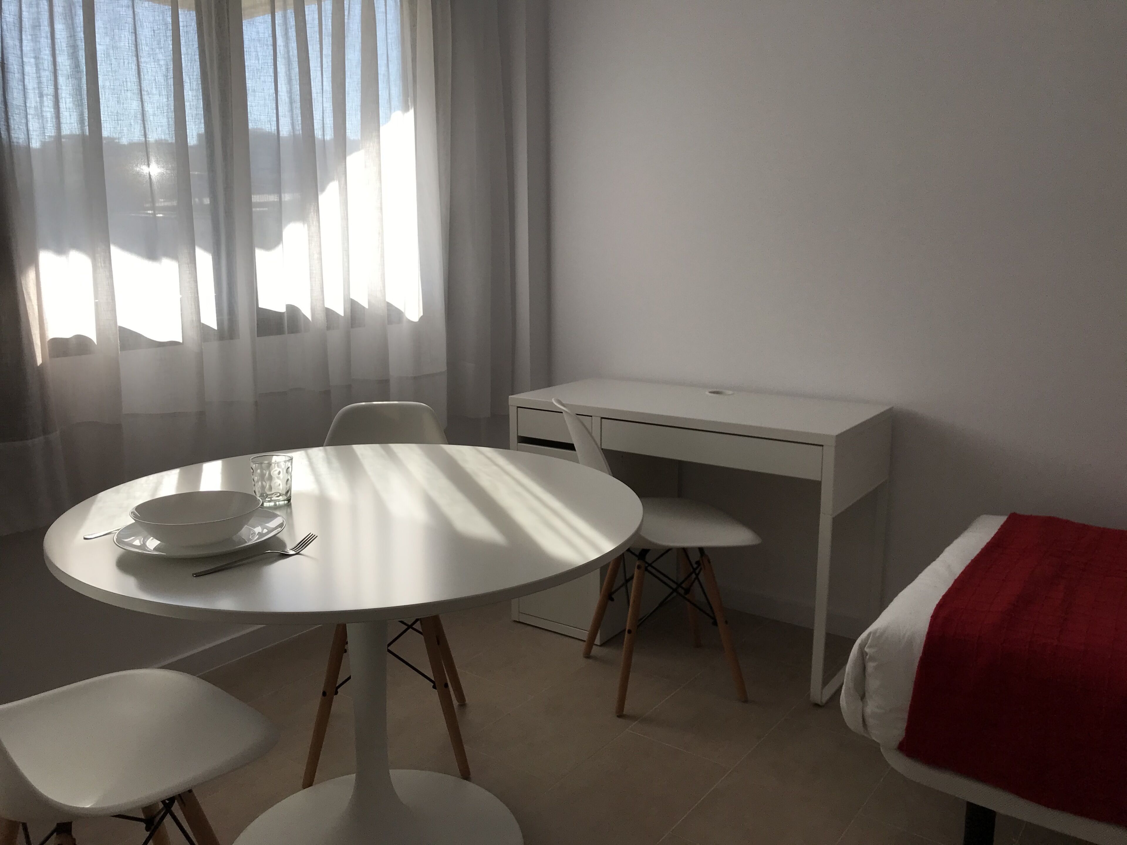 Una habitación con una mesa redonda, dos sillas, un escritorio y una cama con una colcha roja.
