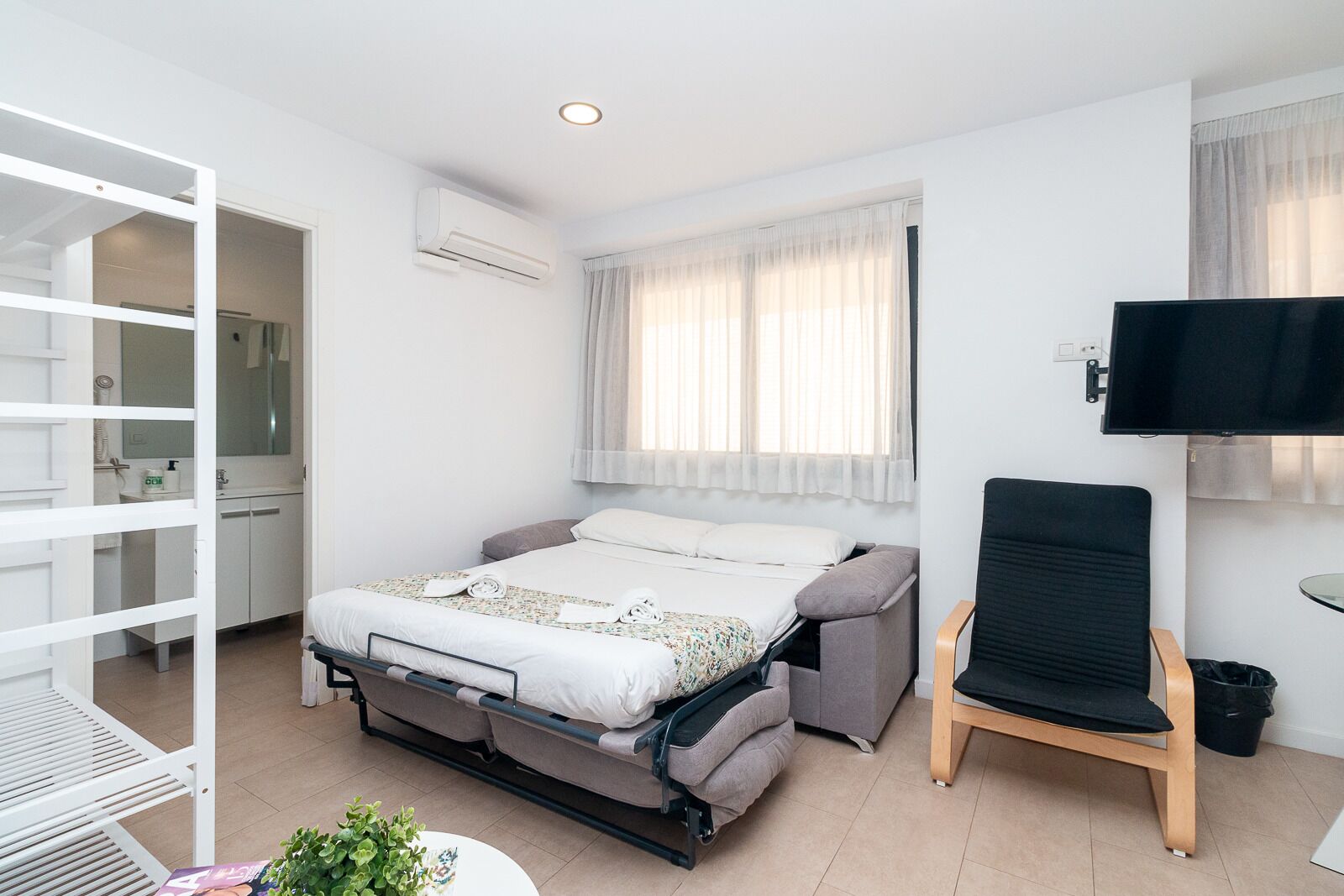 A compact hotel room with a bed, a TV, a chair, and a small table with a plant.