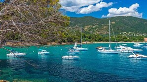 A coastal scene with clear turquoise waters, boats, rocky shore, and a mountainous backdrop. - Studio cabin sea view terrace, internet, air conditioned (Le lavandou)