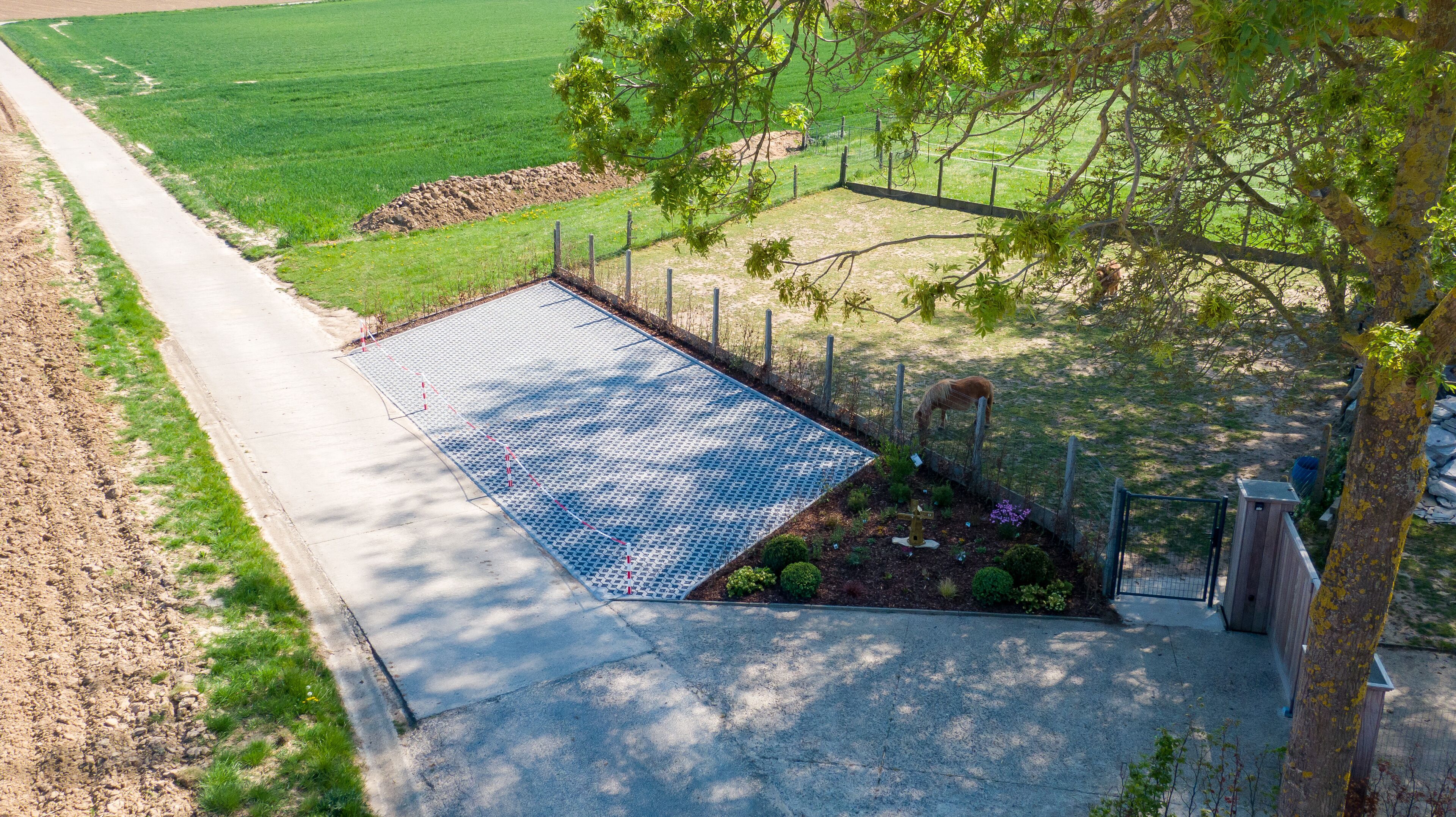 Área pavimentada com superfície estampada, rodeada por cerca e vegetação, com uma árvore em primeiro plano.