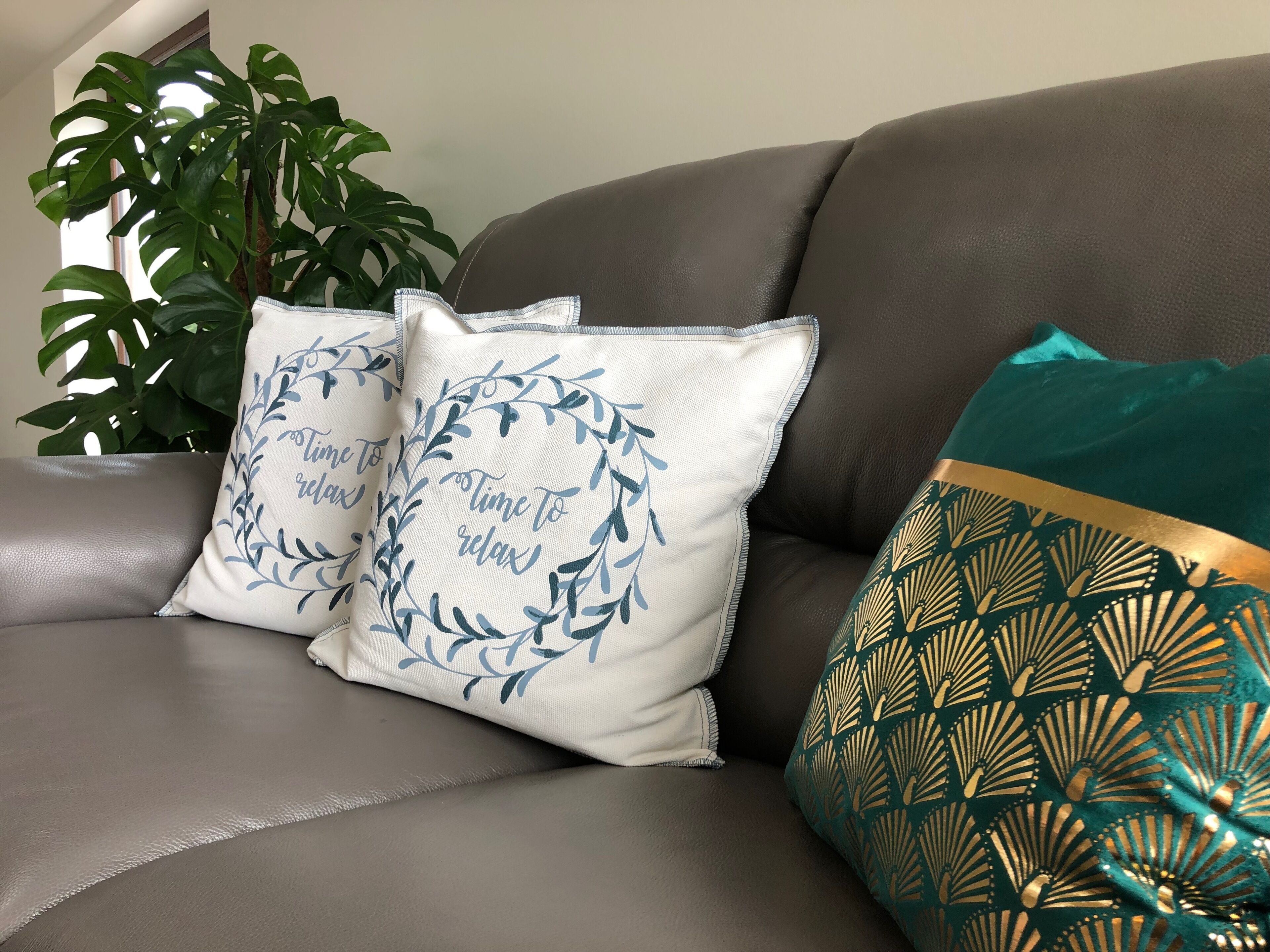 Un canapé en cuir marron agrémenté de trois coussins décoratifs : un avec un motif de feuilles et un texte, un autre avec un motif de feuilles, et un troisième avec un motif doré et turquoise.