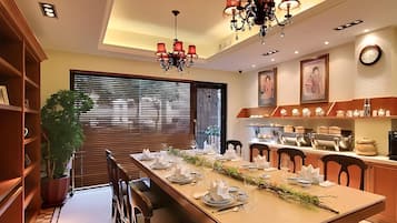 A dining area with a long table set for a meal, wooden chairs, a chandelier, and a kitchen in the background.