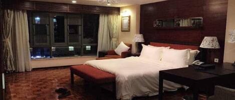 A hotel room with a large bed, a desk, a chair, a window with curtains, and a chandelier.