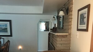 Un salon avec un fauteuil beige, une table à manger avec des chaises, un tapis à motifs, une cheminée en pierre et un coin cuisine avec une fenêtre.