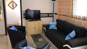Un salon avec un canapé noir, deux fauteuils, une table basse en verre, un téléviseur posé sur un meuble en bois et un rideau à motifs.