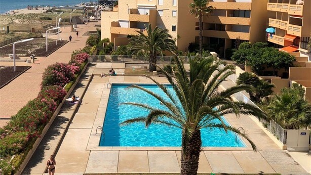 Une piscine avec un palmier au premier plan, entourée d’un espace paysager agrémenté de fleurs, et en arrière-plan, une plage et des bâtiments.