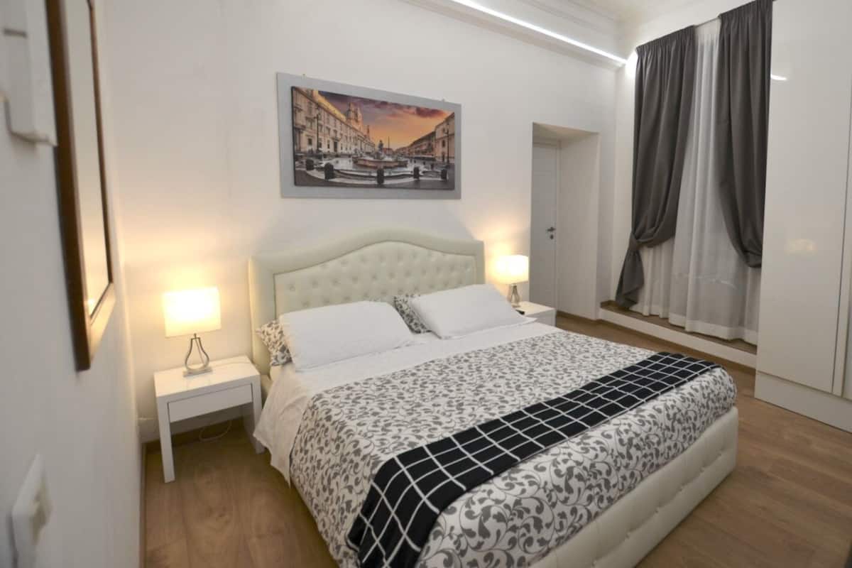 A bedroom with a bed, nightstands, a painting, and a window with curtains.