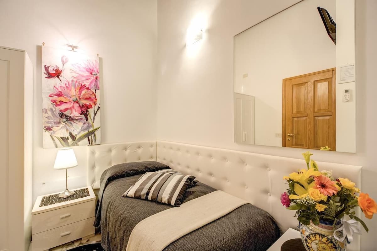 A bedroom with a bed, a nightstand, a lamp, a mirror, and a vase with flowers.