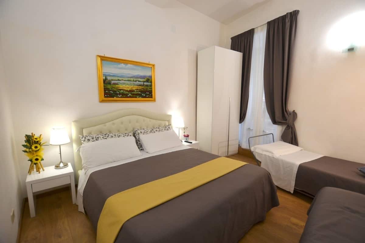 A hotel room with a large bed, a smaller bed, a nightstand with a lamp, a painting on the wall, and a wardrobe.
