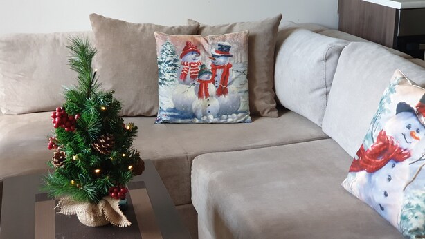 A cozy living room with a beige sofa, a small Christmas tree, and two decorative pillows with winter-themed designs. - Royal Bansko Aparthotel - 1 Bed Sleeps 4 (Bansko)
