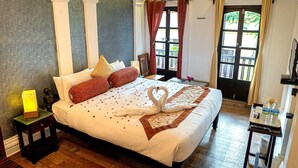 A neatly made bed with a heart-shaped towel arrangement, a bedside table with a lamp, and a window with curtains. - Le Chateau - A Heritage Boutique Hotel (Pondicherry)