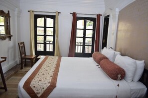 A neatly made bed with white linens and a decorative runner, a wooden chair, and a window with curtains. - Le Chateau - A Heritage Boutique Hotel (Pondicherry)