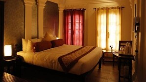 A hotel room with a large bed, a nightstand, a small table, and a chair. - Le Chateau - A Heritage Boutique Hotel (Pondicherry)